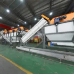 this image depict an industrial plastic recycling washing system or recycling plant. The large machinery with conveyor belts and sorting components is typical of equipment used to wash, sort and process plastic waste materials as part of the plastic recycling process. These systems are designed to efficiently handle large volumes of plastic items or bales, separating them by type, removing contaminants through washing/rinsing stages, and preparing the cleaned plastic materials for further recycling and reprocessing into new plastic products or raw materials. The orange and gray coloring of the machinery is also characteristic of many plastic recycling facilities and systems.
