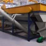 part of a plastic recycling facility. In such a system, the yellow hopper could be used to collect and funnel plastic waste into the machine. The segmented chute would then guide the plastic through various stages of sorting, shredding, washing, and possibly even pelletizing, depending on the complexity of the facility. The grey metal bin might be where the sorted and processed materials are collected before they are moved to the next phase, which could involve further refinement or packaging for transport. The green dumpster on the left could be for non-recyclable waste that is separated out during the process. This setup is essential in the recycling loop, helping to reduce plastic waste by converting it into reusable materials.